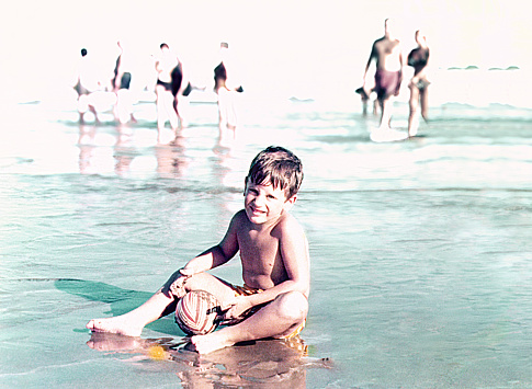 Vintage image of a young boy sitting close to the water, at the beach, and playing with a ball. Vintage photo of the seventies of the 20th century.