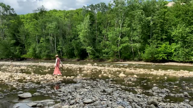 Retro styled young woman in straw boater hat and long dress walking on river rocks near forest. Romantic girl. Natural scenic landscape. Summer vacation. Vintage aesthetic lifestyle. 4K video