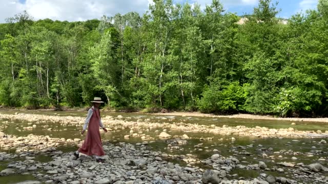 Retro styled young woman in straw boater hat and long dress walking on river rocks near forest. Romantic girl. Natural scenic landscape. Summer vacation. Vintage aesthetic lifestyle. 4K video