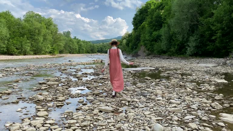 Retro styled young woman in straw boater hat and long dress walking on river rocks near forest. Romantic girl. Natural scenic landscape. Summer vacation. Vintage aesthetic lifestyle. 4K video