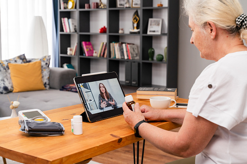 back view hoary old woman in sit at table at home look at digital tablet screen answer question about health talks with therapist by video conference call application. Remote medical consultation concept