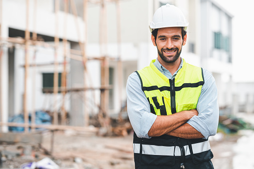 Happy caucasian man construction site manager smiling with confident standing arms crossed wearing safety vest and helmet at construction site.