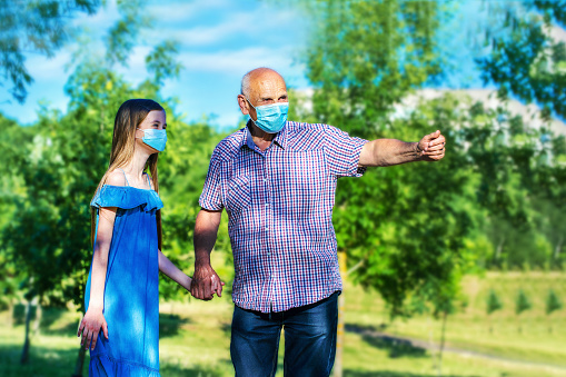 Grandfather with granddaughter wearing medical face mask walking at urban summer park.