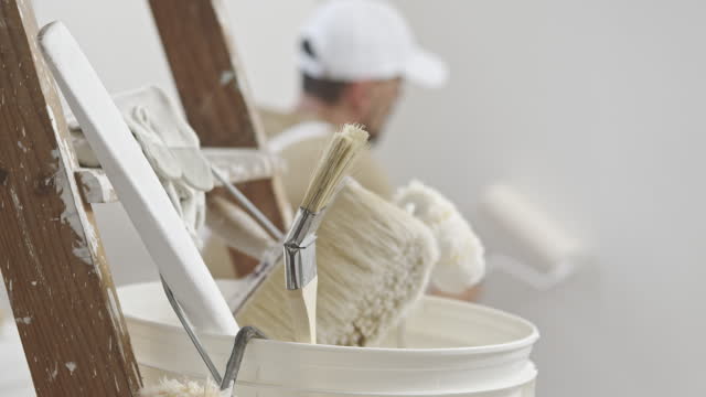 hands of house painter man decorator work of home to renovate, using roller paint and holding white bucket, a wooden ladder with paint brushes as background, close-up