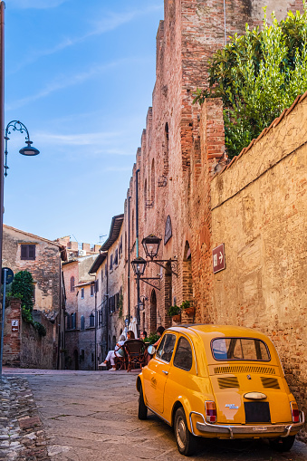 Vintage yellow Fiat 500 parked in an alley in the old town of Certaldo, a town in the Metropolitan City of Florence. People sitting at a bar outdoor tables