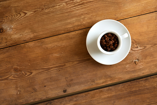 Overhead view of a white cup filled with coffee beans with saucer on wooden table with seamless pattern