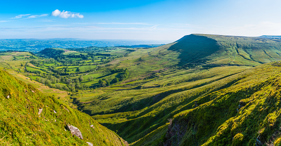 Panoramic view over the escarpment of the Black Mountains and Gospel Pass descending to the picturesque patchwork landscape of pasture, meadows and farm fields under blue summer skies, Wales, UK.
