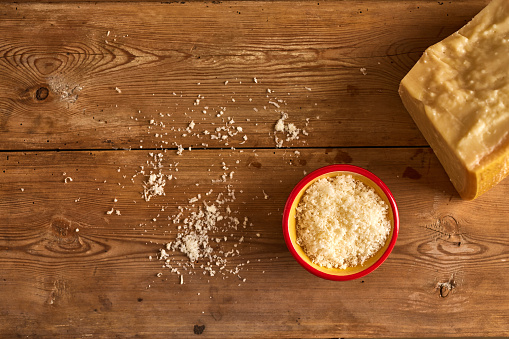 Overhead view of finely grated cheese in a bowl with block with cheese sprinkled on wooden table