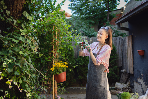 A smiling silver-haired woman stands holding a watering can and wearing gardening clothes in her back yard, near a flowering clematis