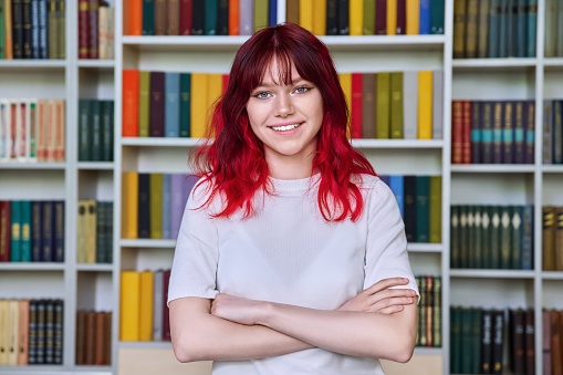 Confident teenage female student with crossed arms looking at camera, portrait in library. Teenage boy with red hair, education, knowledge, back to college concept