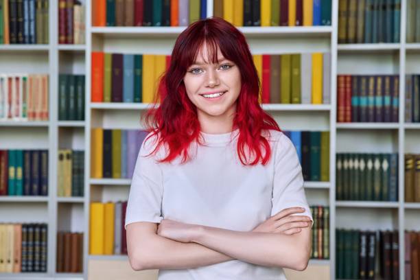 Confident teenage female student with crossed arms looking at camera, portrait in library stock photo