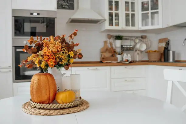 Photo of Still-life. Yellow, orange flowers in a vase, pumpkins and candles on a golden tray on a white table in a home kitchen interior. A cozy autumn concept.