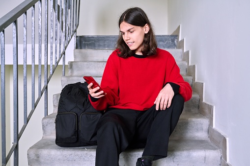 Young male sitting on stairs using smartphone. Handsome teenage guy student with backpack with phone in his hands. Youth, lifestyle, network, leisure, network, leisure, young people concept