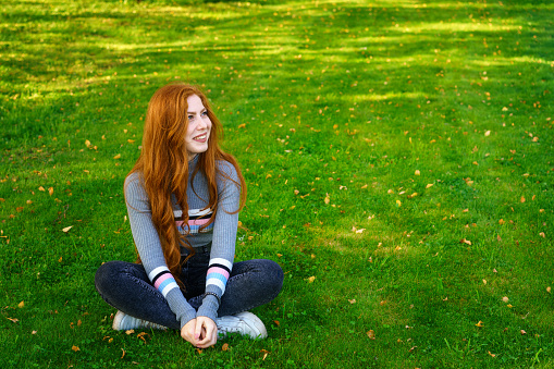 happy young woman of Caucasian ethnicity, with long red hair sits in casual clothes in the park on green grass smiling and looking to the side on a sunny day