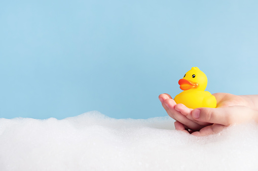 Child hands holding yellow rubber duck in bubble bath on pastel blue background. Copy space