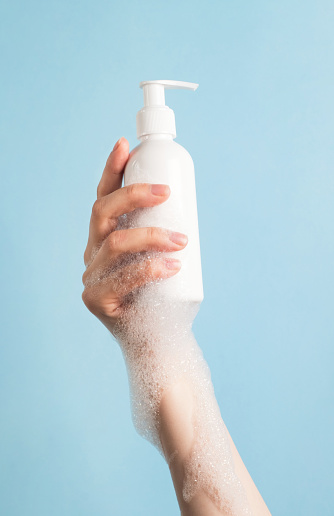 Womans hand holding cosmetics bottle with foam bubbles on pastel blue background. Vertical, close up