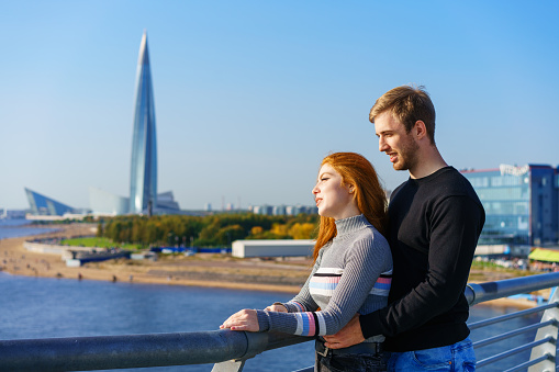 A guy and a girl of Caucasian ethnicity stand together on a river bridge on a sunny day with a city view