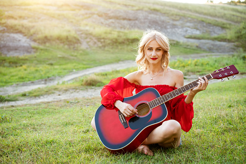 The woman in red is sitting on the green grass with a red guitar. Girl musician plays the guitar outdoors. Blonde caucasian appearance