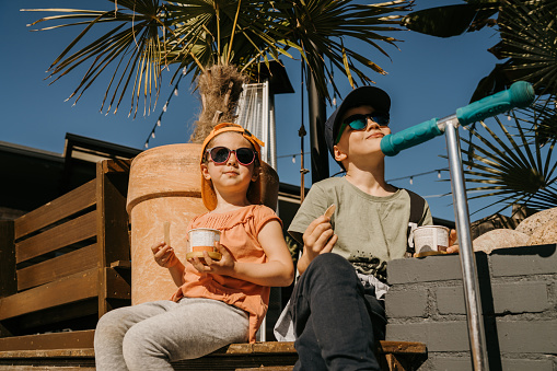 Little brother and sister taking a break eating ice cream after riding a push scooters together.