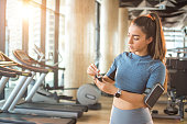 Portrait of beautiful fit girl preparing for treadmill. Sportswoman taking wireless earphones connected to smartphone on her armband and her smartwatch. Technology and sports activity concept