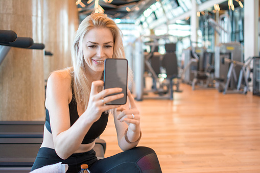 Blonde woman in sportswear sitting on treadmill machine during exercise break and using smart phone at gym