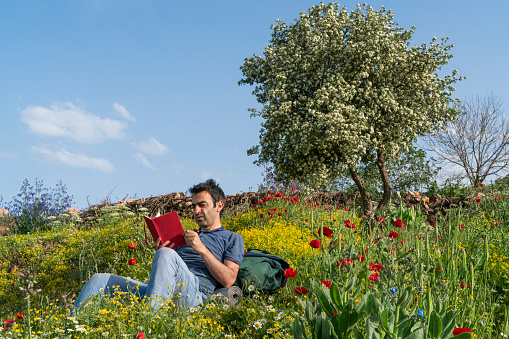 The man is sitting among the wildflowers and reading a book. The man is resting, leaning on his backpack. There is a blooming hawthorn tree right behind it. Taken with a full-frame camera in sunny weather.