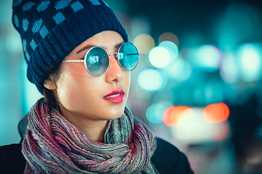 In this outdoor image with copy space, an Asian/Indian, single young woman enjoys her leisure by walking at night in a city street. She wears a warm cap, muffler coat, and sunglasses.