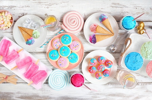 Pastel summer sweets table scene. Collection of ice cream, popsicles, cookies and treats. Overhead view over a rustic white wood background.