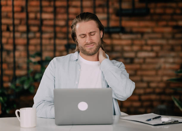 istock neck pain while working. Young guy holding his neck while working with a laptop in the office 1401023257