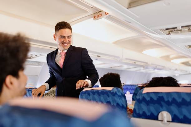 Male Cabin Crew member walks down the aisle checking on passengers Male flight attendant checking on the passengers before take off crew stock pictures, royalty-free photos & images