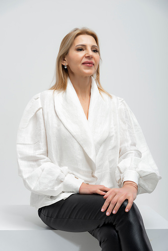 The mature and beautiful photomodel wearing a business white suit shorts poses artistically in the studio with a white background. Photo shoots for the clothing industry, which is a textile product for e-commerce. Image work. Cover images. A brunette, wheat-skinned mature woman with blonde hair.