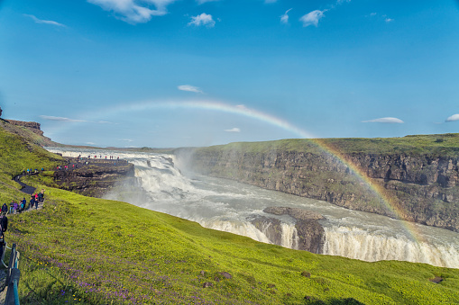 Rainbow over Gullfoss waterfall on Hvítá river and line of incidental unrecognizable tourists on footpath. Iceland