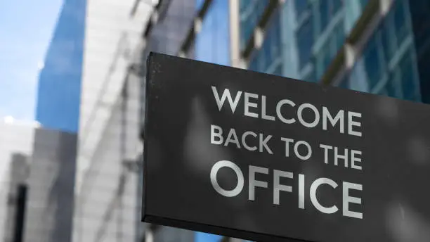 Welcome back to the Office on a black city-center sign in front of a modern office building