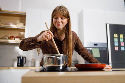 Cheerful young woman cooking and stirring food in saucepan at the kitchen