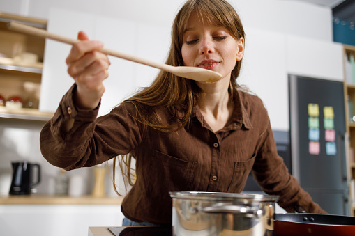 Pretty girl tasting food with wooden spoon while cooking