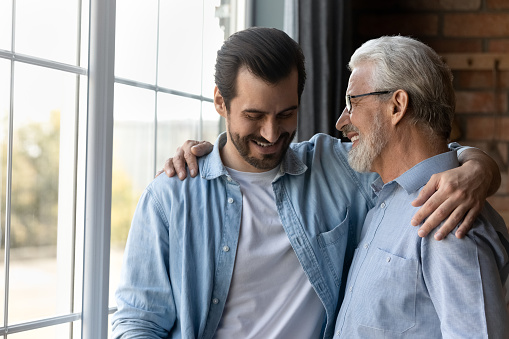 Close up happy mature father and grownup son hugging cuddling enjoying tender moment, standing at home, smiling young man with senior dad in glasses having fun, two generations good relationship