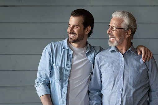 Happy two generations of men hugging, looking in distance, hugging, standing on grey wooden wall background, smiling senior father in glasses and grownup son dreaming, family unity and bonding