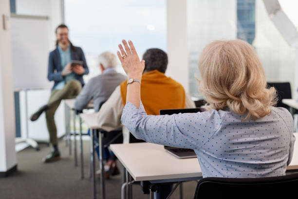 A senior woman is asking for an explanation in the computer course while sitting with elderly students in the classroom. stock photo