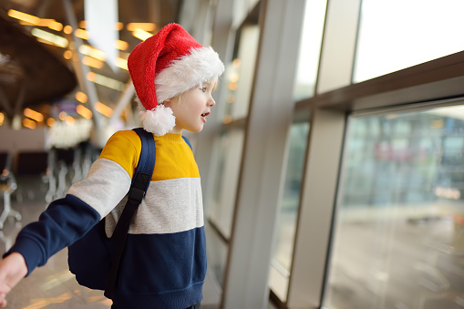 Little child wearing Santa Claus hat on background of an international airport. Preschooler boy is waiting for a merry Christmas.. Family trip for winter holidays. Kids vacation. Baby admiring airplan