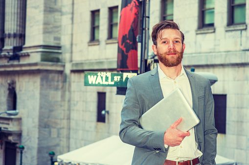 American Businessman with beard, mustache traveling, working in New York, wearing cadet blue suit, white undershirt, carrying laptop computer, standing on Wall Street by street sign, looking forward.