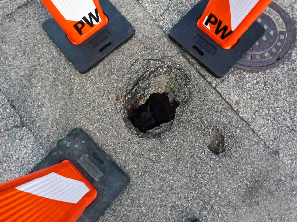 Large, deep pothole in the street surrounded by orange and white barricades. View from directly above looking down into the hole in the street.