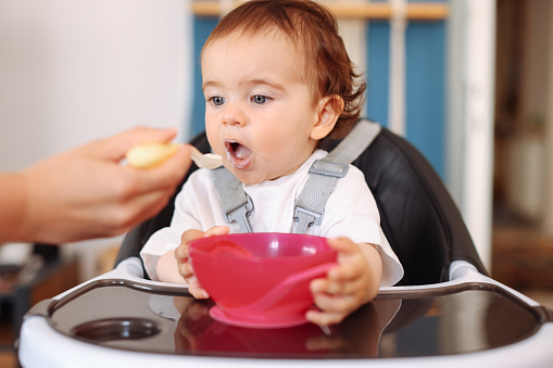 An adorable baby in a high chair laughs at her mother as he spoon feeds his. He pretends the spoon is a moving train using sound effects