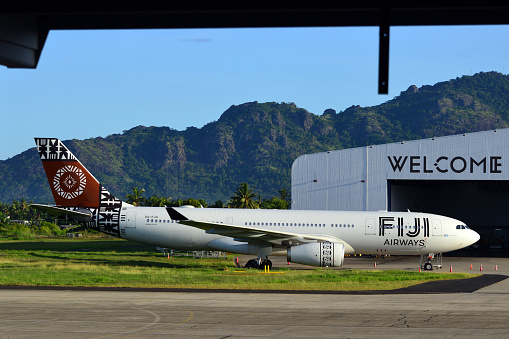 Nadi, Viti Levu island, Fiji: Fiji Airways Airbus A330-243 registration DQ-FJO, named Island of Beqa - Nadi International Airport - 'welcome' on hangar façade, tropical forest and mountains in the background.