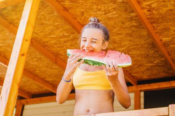 육즙이 많은 수박을 먹는 공원의 귀여운 소녀. 여름 테라스에있는 소녀는 붉은 수박의 큰 조각을 먹고 미소를 짓습니다. 음식 자연. 사랑스러운 금발 소녀는 야외에서 수박 조각을 먹습니다. - watermelon missing bite melon grass 뉴스 사진 이미지