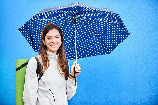 Portrait of a beautiful young woman with yoga mat and umbrella standing against blue wall