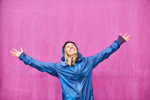 Portrait of cheerful young woman with her raincoat enjoying in heavy rain against purple background