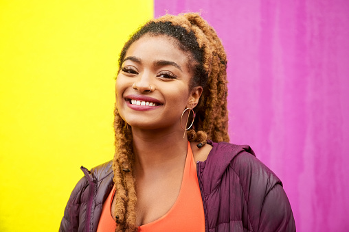 Portrait of stylish young african woman with dreadlock standing against yellow and purple wall looking at camera and smiling