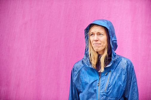 Portrait of a woman wearing raincoat looking upset with heavy rain while standing against purple background