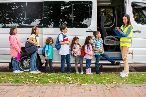 Happy Latin American teacher in charge of a group of children boarding the school bus and writing their names on a clipboard