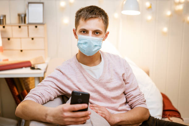 istock a young man sits on a sofa wearing a protective mask with a phone in his hand 1401104928